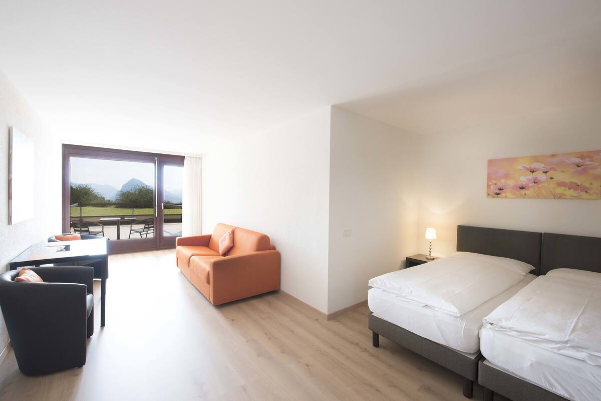 A modern hotel room with a bed, a sofa, a desk, and a view of the outdoors.