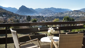 A balcony with wicker chairs and a table, overlooking a mountainous landscape with a town below. - Centro Cadro Panoramica (Lugano)