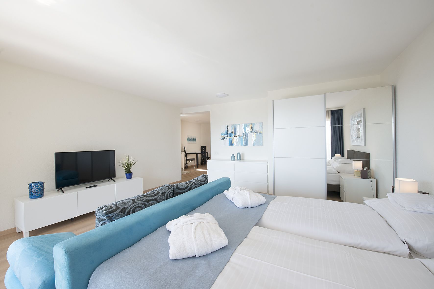 A modern hotel room with a large bed, a TV, a sofa, and a dining area visible in the background.