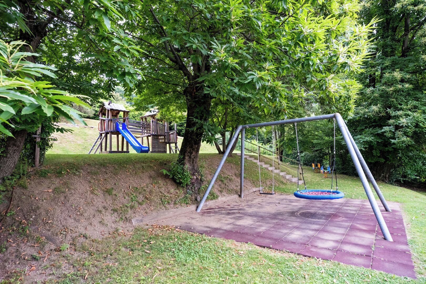 Un terrain de jeux avec des balançoires et un toboggan, entouré d’arbres et d’herbe.