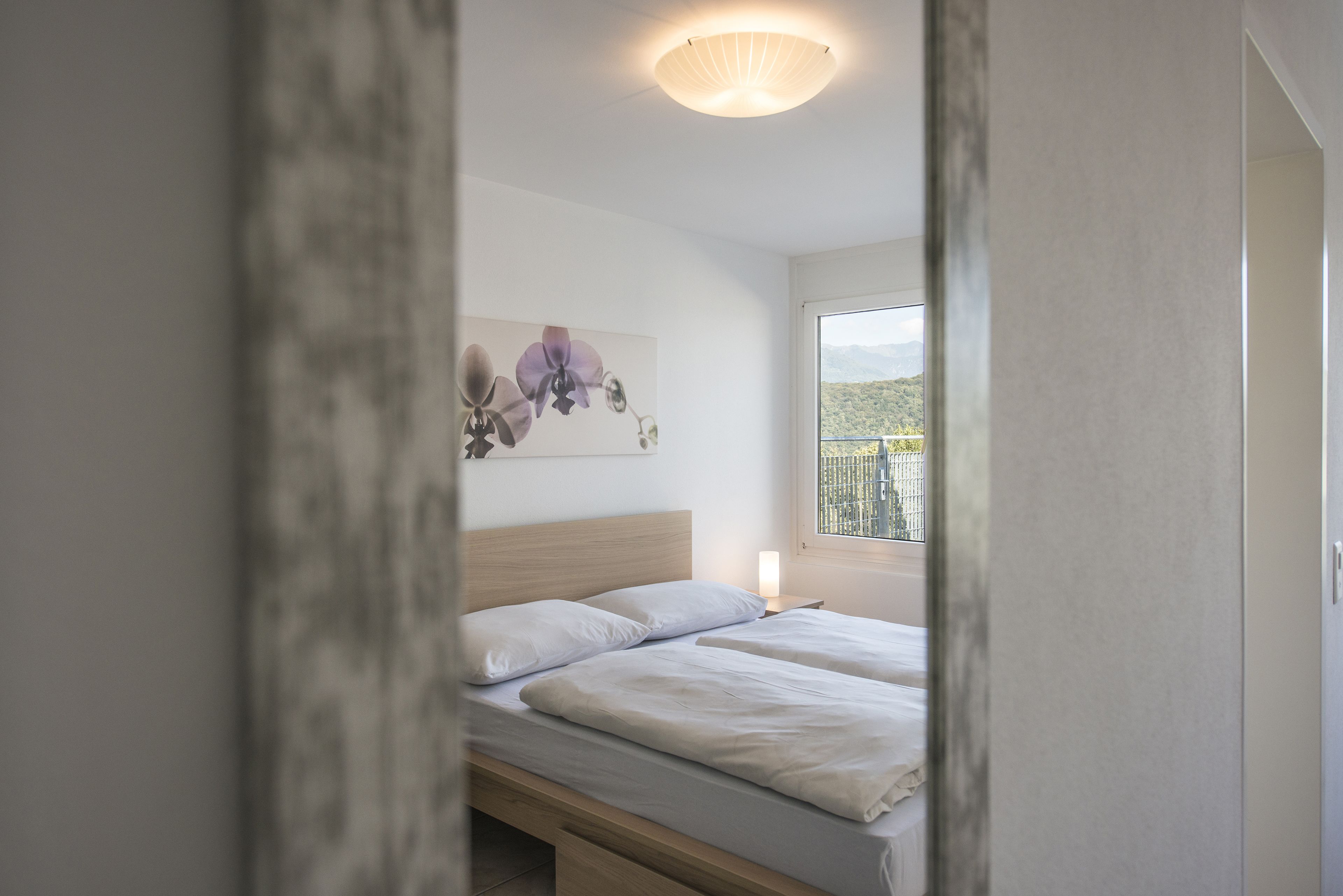 A bedroom with a bed, a nightstand, a wall-mounted light, and a window with a view of the outdoors.