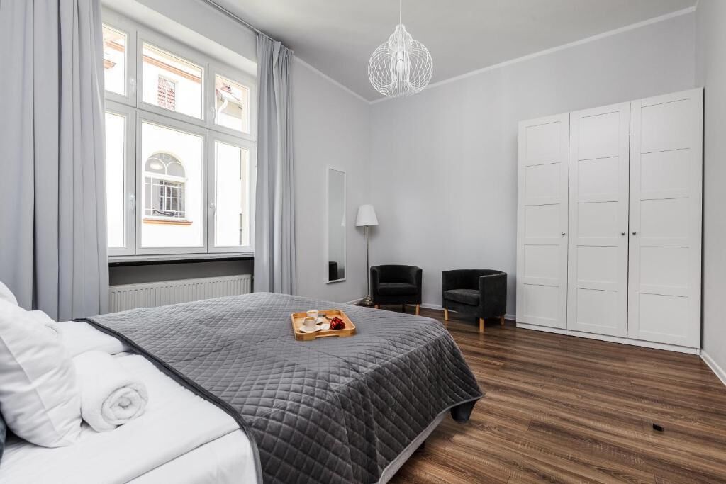A hotel room with a bed, a window with curtains, a chandelier, a wardrobe, and a small table with a tray.