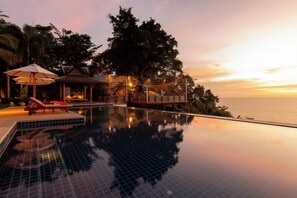 Ein Pool ohne Ende mit freiem Blick auf den Ozean bei Sonnenuntergang, umgeben von Palmen und einem Pavillon.
