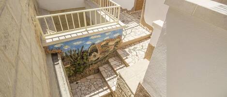 Un escalier avec une rampe blanche, un mur bleu et beige orné d’une fresque, et une porte avec un volet en bois.