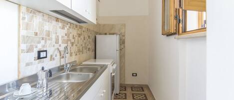A narrow kitchen with a tiled backsplash, stainless steel countertops, and a refrigerator.