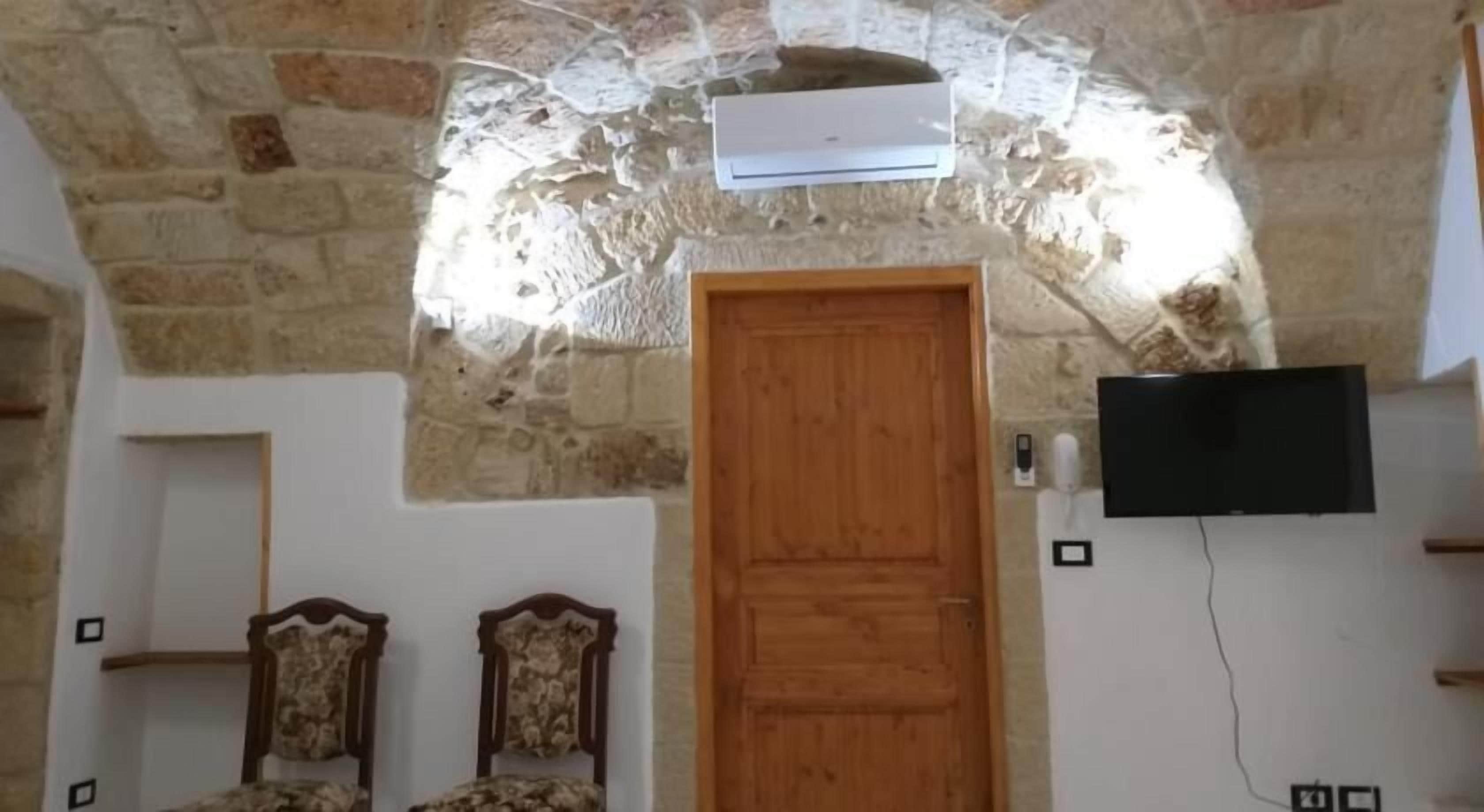 Ein Zimmer mit Steindecke, einer Holztür, zwei Holzstühlen und einem an der Wand befestigten Flachbildfernseher.