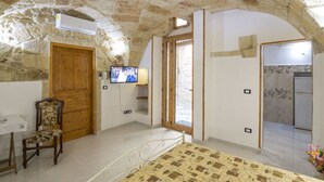 A bedroom with a stone ceiling, a wooden door, a bed with a patterned bedspread, a chair, a television, and a bathroom visible through an open door. - Dimora Corte Antica Scorrano (Scorrano)