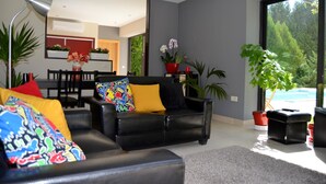 Een moderne woonkamer met een zwarte leren bank, kleurrijke kussens en een groot raam met uitzicht op een zwembad en groen.