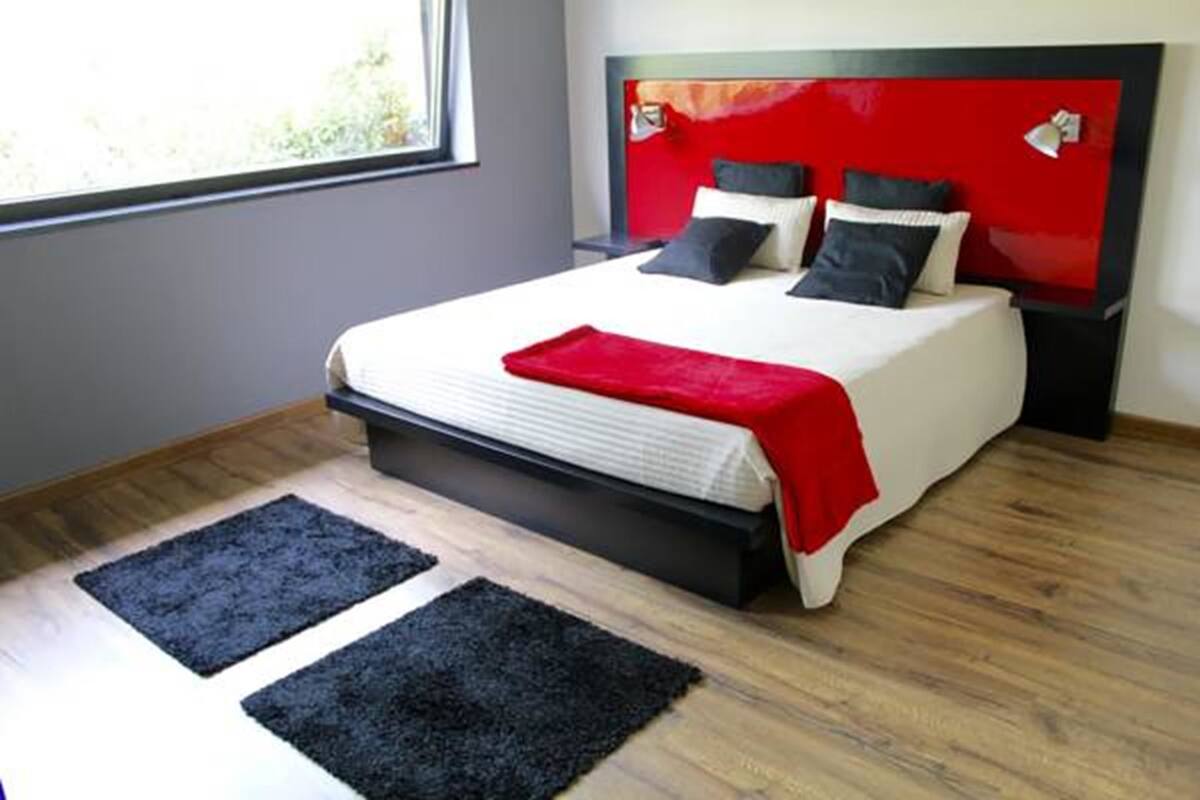 Une chambre moderne avec une tête de lit rouge, un lit recouvert de linge de lit blanc et deux tapis noirs sur le sol.