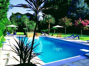 A swimming pool with lounge chairs and umbrellas, surrounded by greenery and a house in the background.