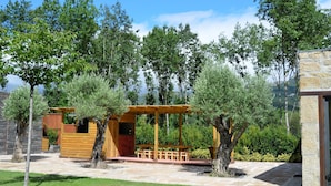 Een houten pergola met een geïntegreerd barbecuegedeelte, omringd door bomen en een stenen pad.