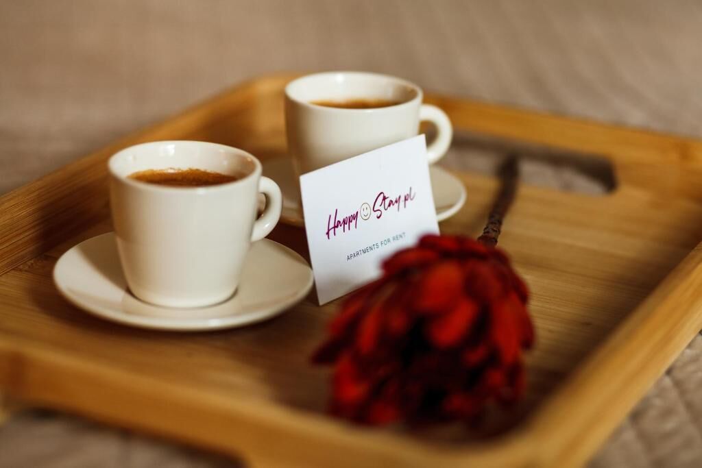 Two white cups of coffee on a wooden tray with a red rose and a card that reads 'Happy Stay'.