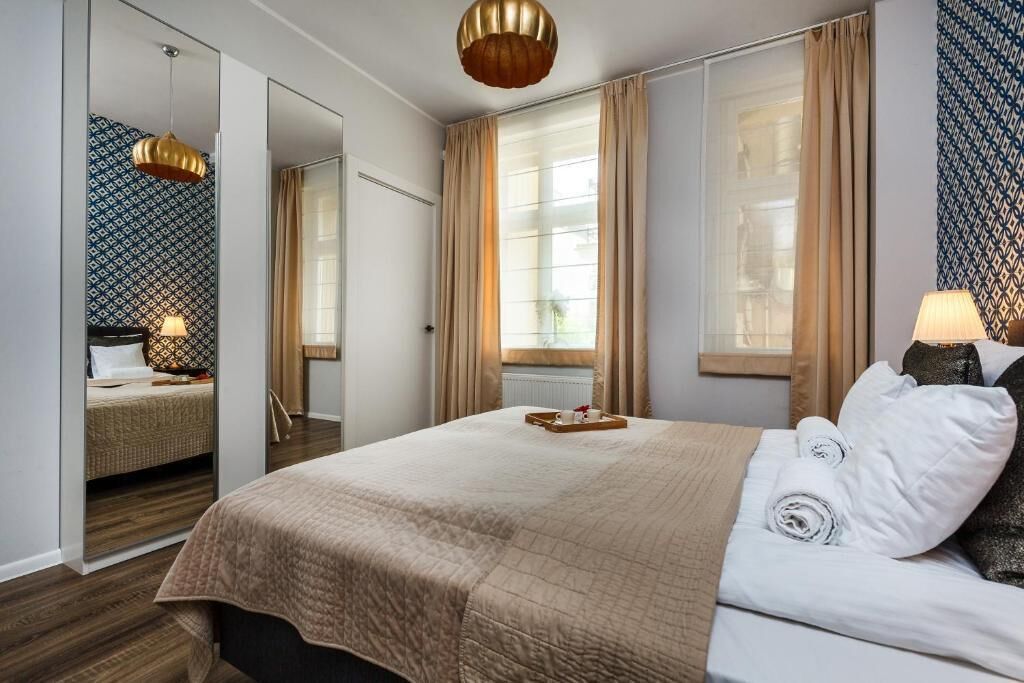 A hotel room with a large bed, a mirror, a window with curtains, and a bedside table with a lamp.