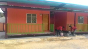A red building with a green railing and a door. Two people are painting the ground. - SiBamboo 2 (Chiang Rai)