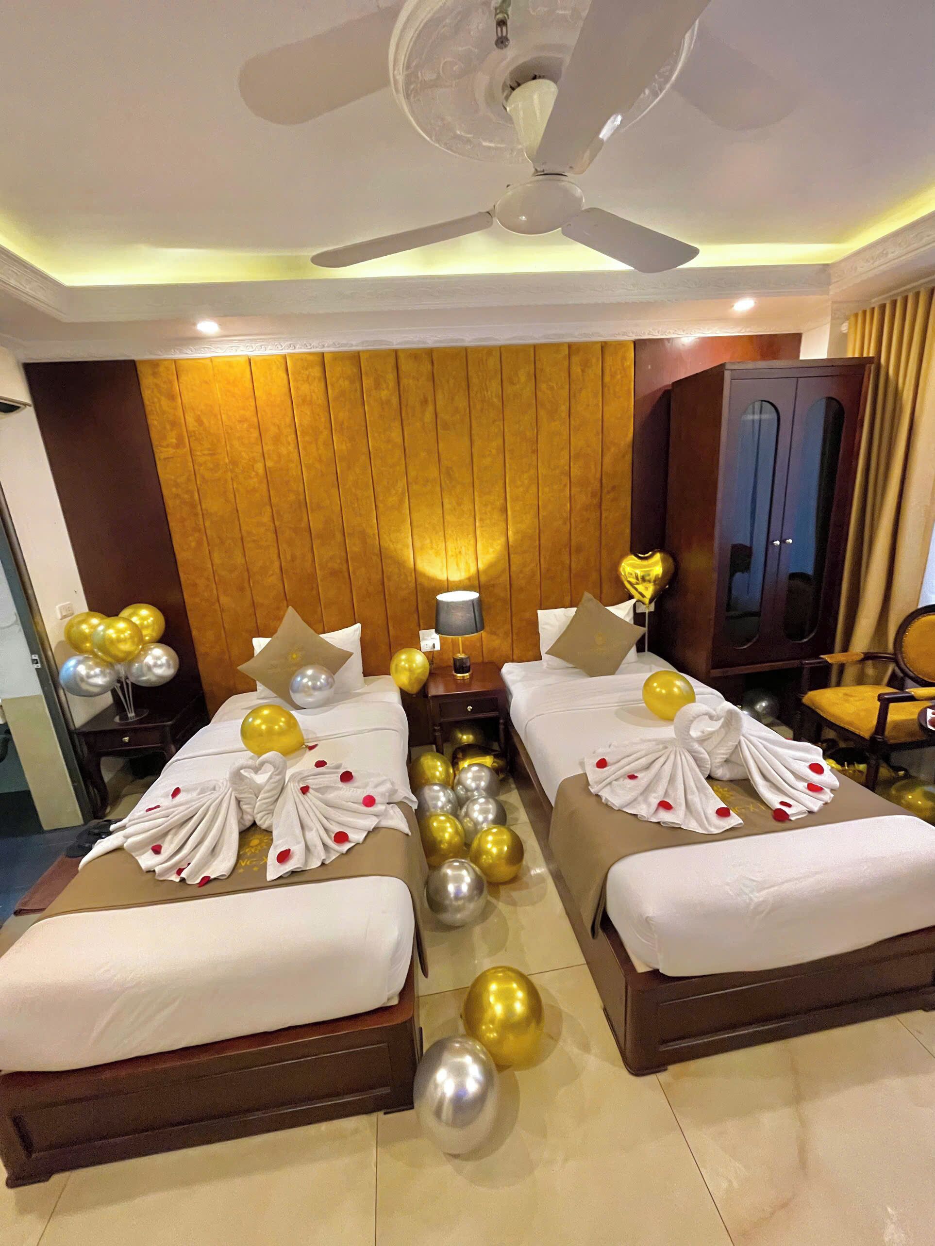 A hotel room with two beds, a ceiling fan, a wooden headboard, a wardrobe, and balloons.