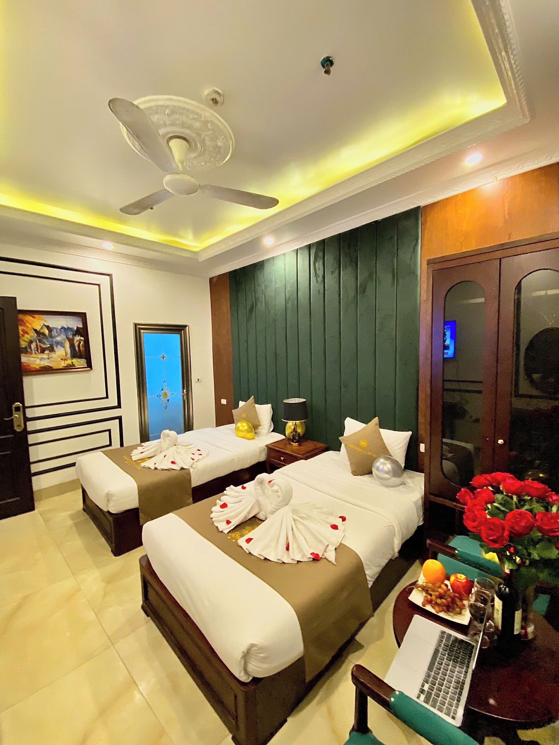 A hotel room with two beds, a desk with a laptop, a vase of red roses, and a door with a mirror.