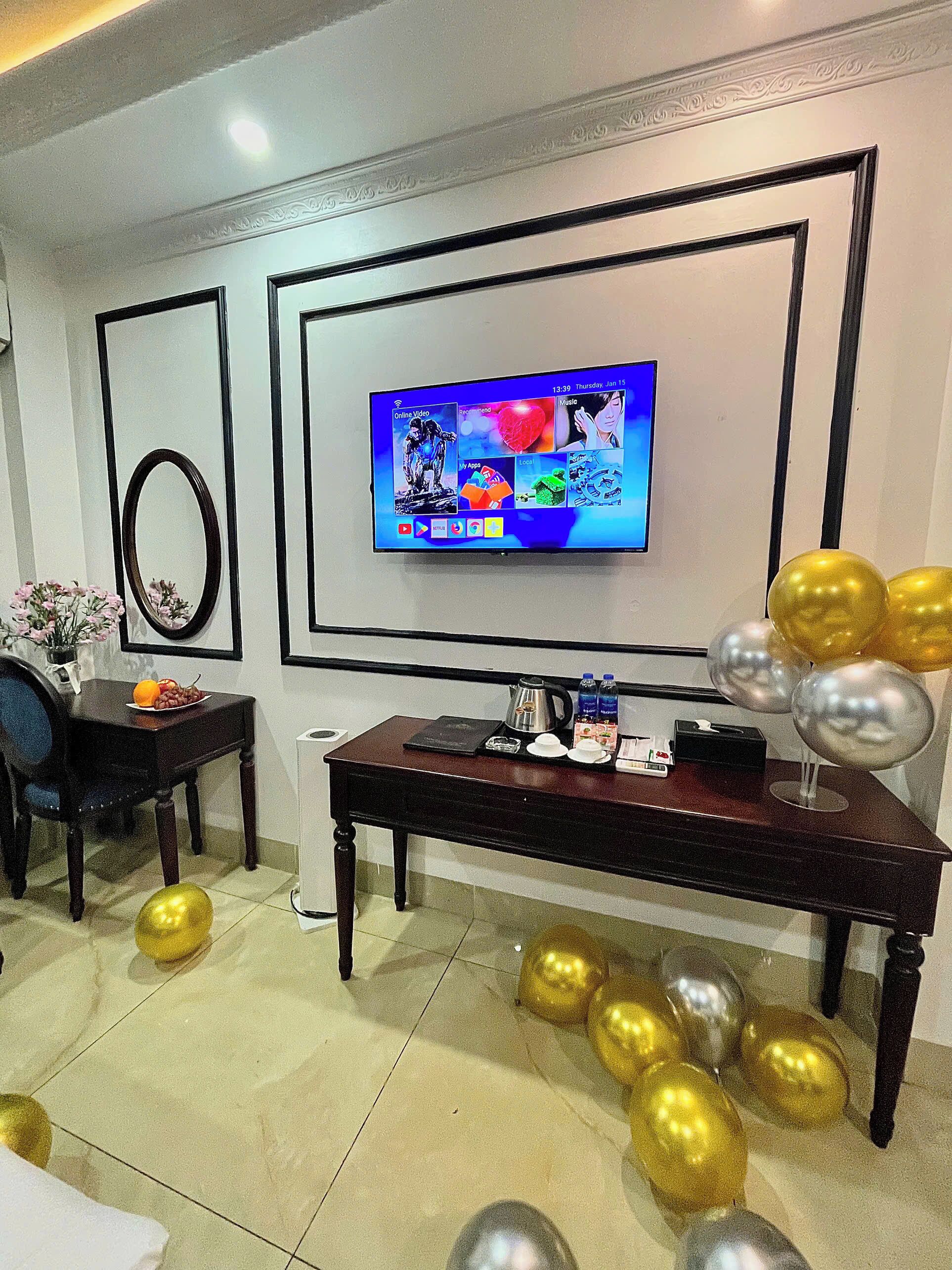 Quarto moderno com televisão de tela plana na parede, uma mesa de jantar com cadeiras e um arranjo decorativo de balões.
