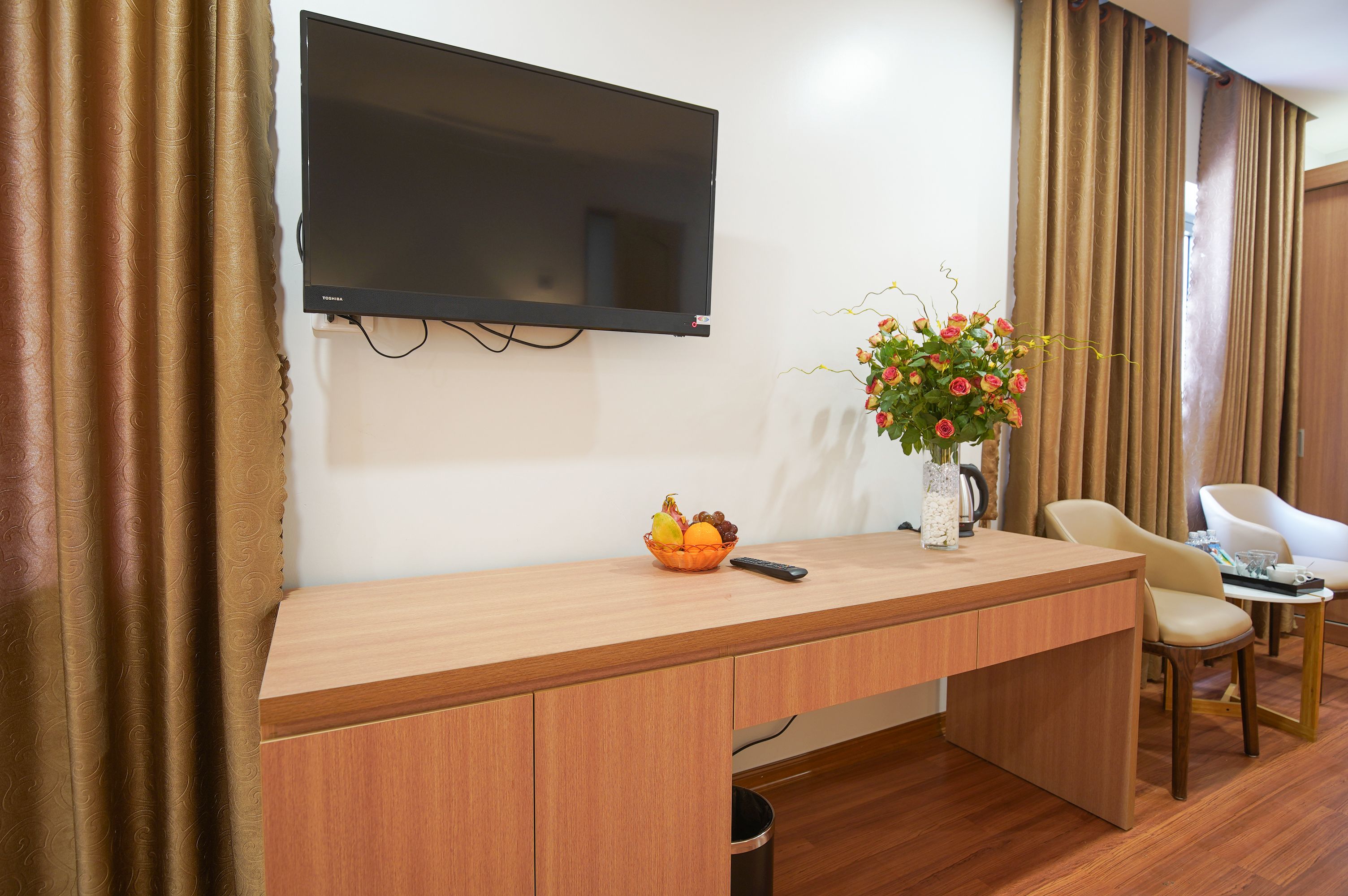 A hotel room with a wooden desk, a flat-screen TV mounted on the wall, a vase with flowers, a fruit bowl, and a remote control.