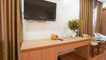 Une chambre d’hôtel avec un bureau en bois, un téléviseur à écran plat fixé au mur, un vase avec des fleurs, un panier de fruits et une télécommande.