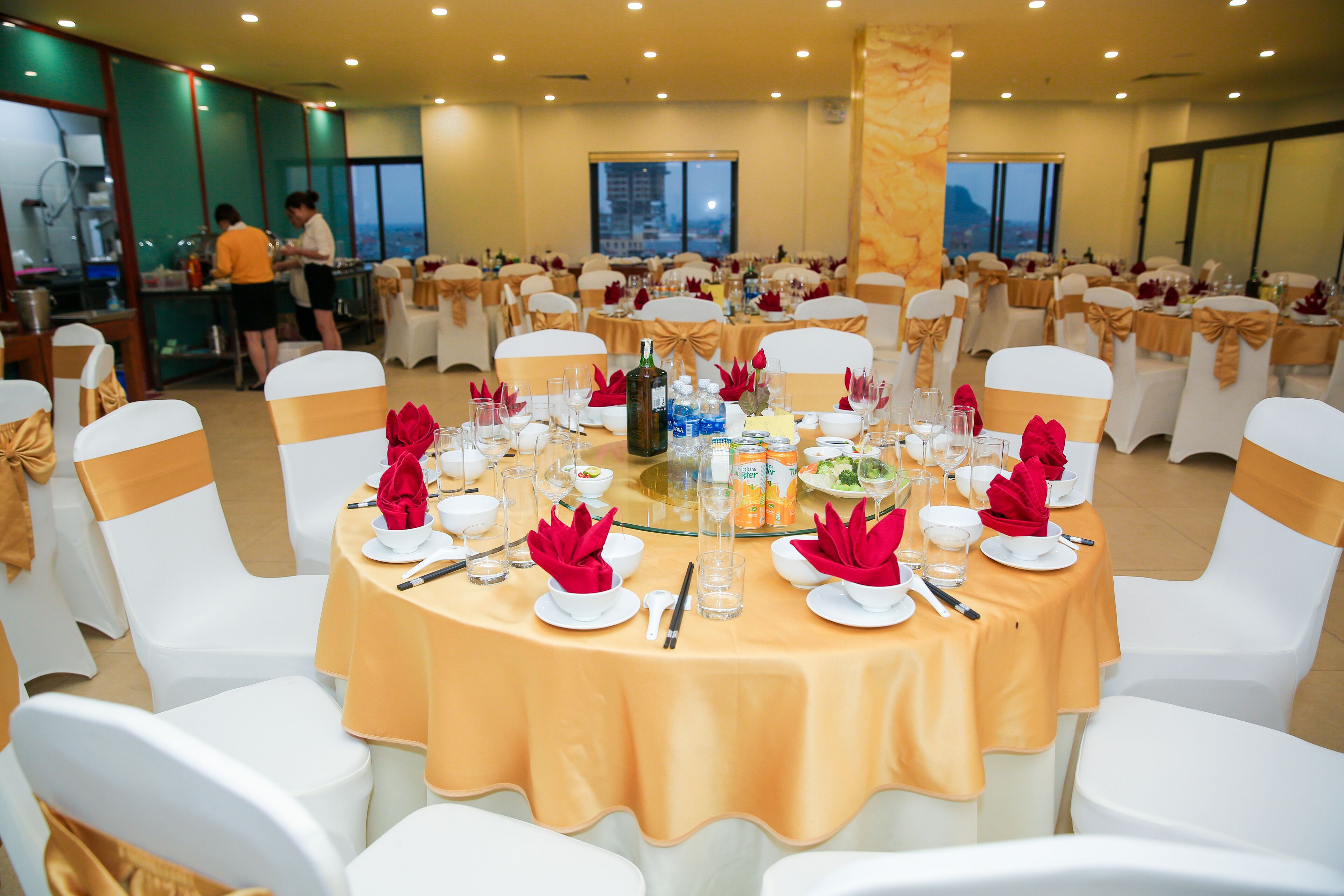 A banquet hall with round tables set for a formal event, featuring white chairs with golden sashes, and a view of the city through large windows.