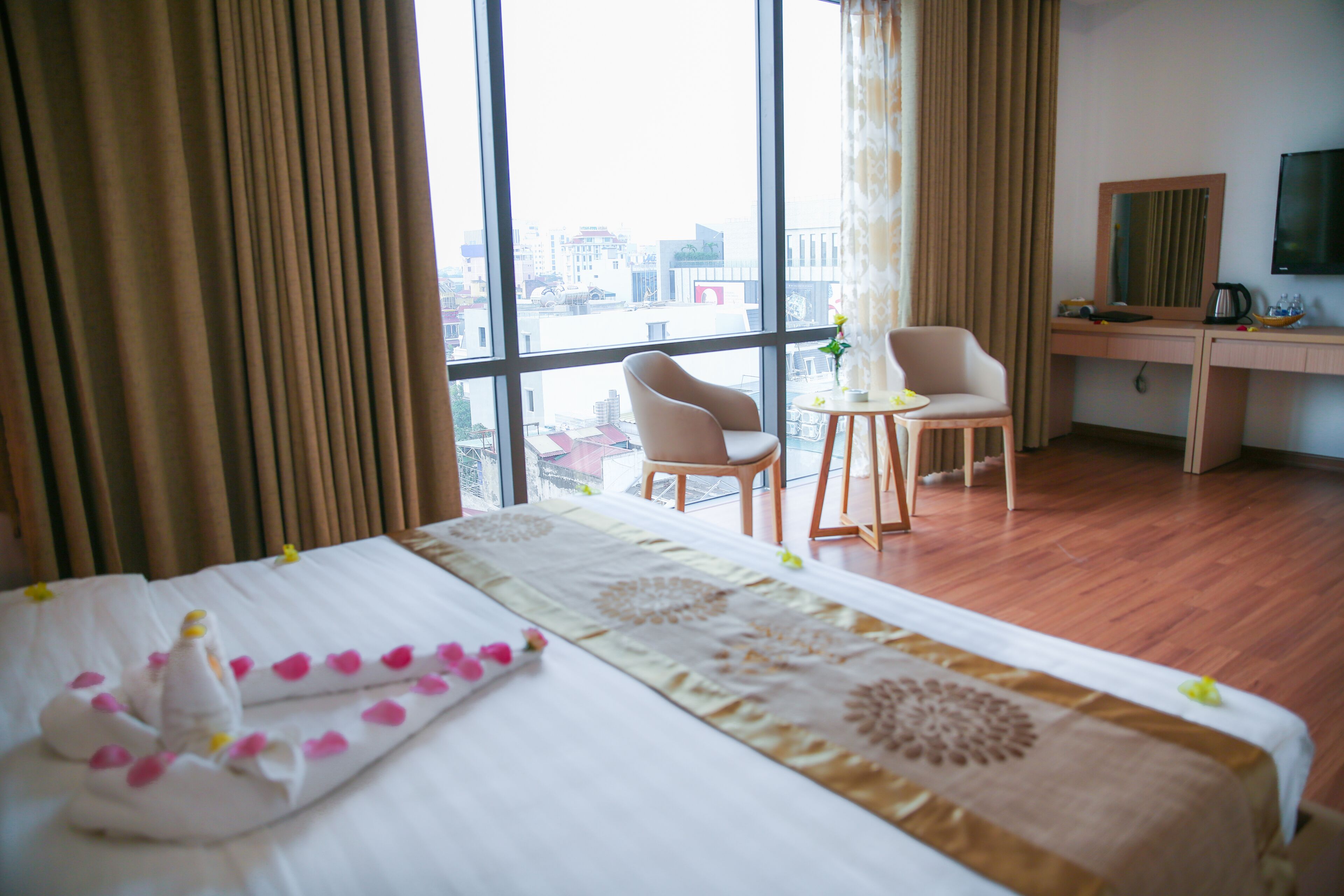 A hotel room with a large bed, two chairs, a desk, and a view of the city through large windows.