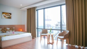 Habitación de hotel con una cama grande, dos sillas, una mesita y vista a una obra en construcción a través de amplias ventanas.