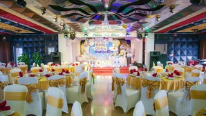 Un salón de banquetes con filas de mesas y sillas, un escenario con un banderín y un techo con luces decorativas.