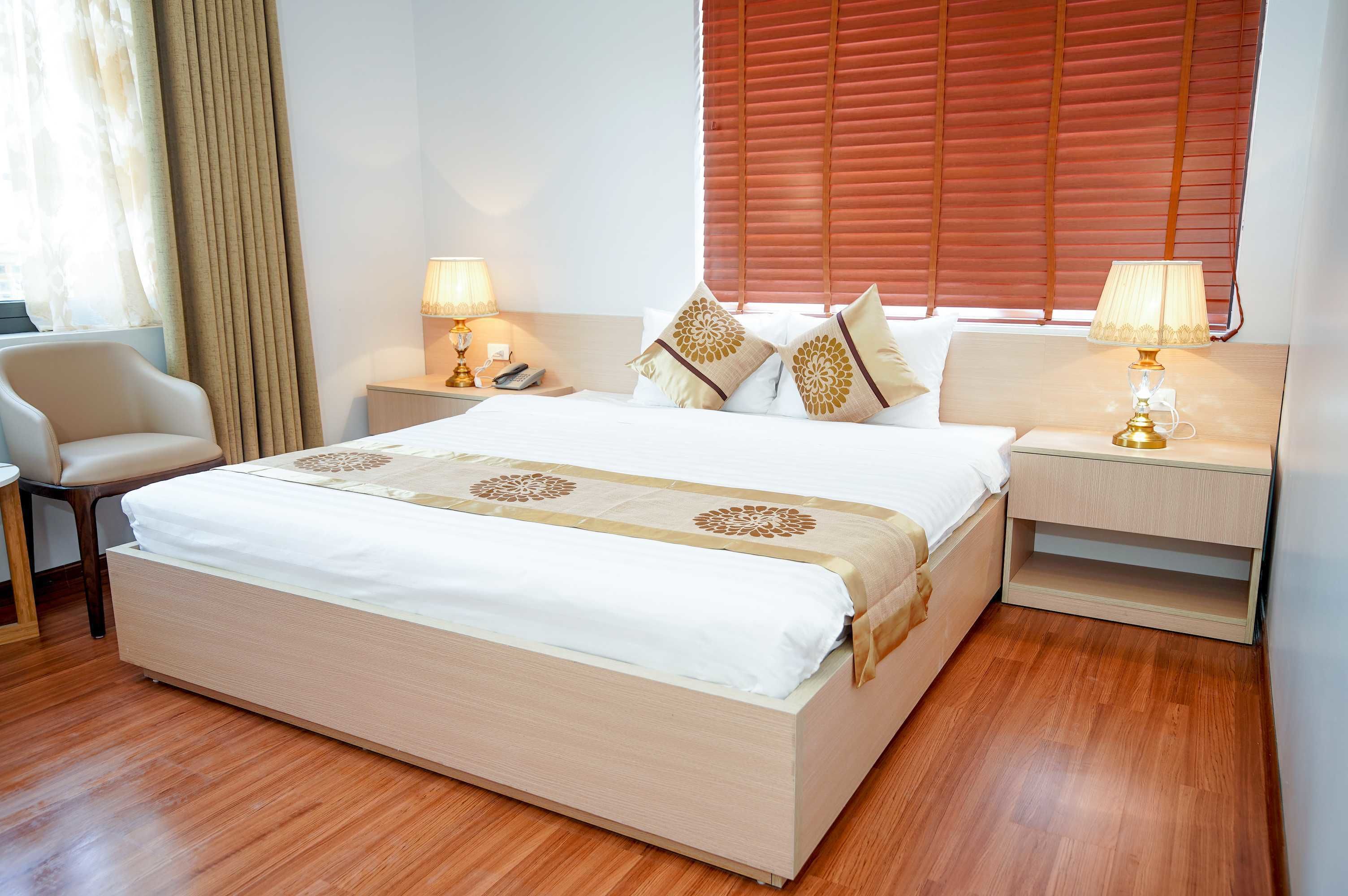 A neatly made bed with a white and beige bedspread, flanked by two bedside tables with lamps, a chair, and a window with blinds.
