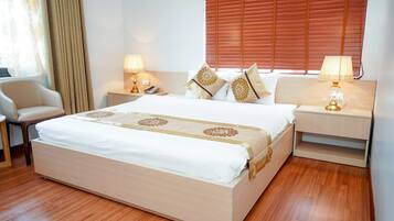 A neatly made bed with a white and beige bedspread, flanked by two bedside tables with lamps, a chair, and a window with blinds.