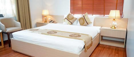 A neatly made bed with a white and beige bedspread, flanked by two bedside tables with lamps, a chair, and a window with blinds.