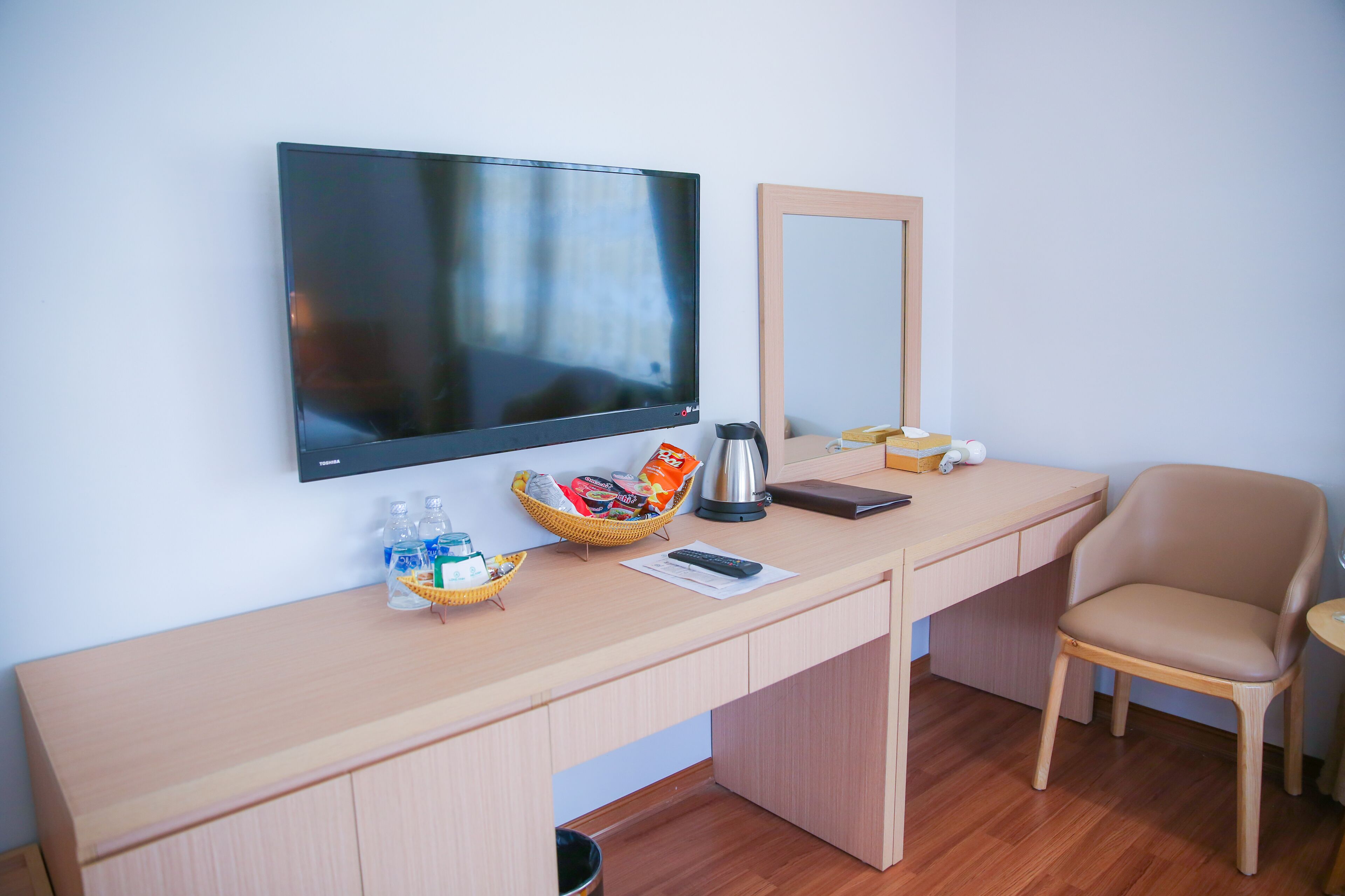 A hotel room with a wooden desk, a television mounted on the wall, a chair, and a small table with a kettle and a basket of snacks.
