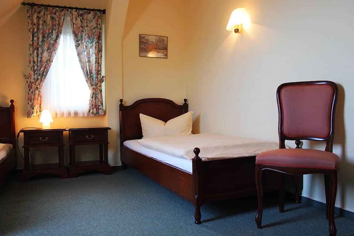 A bedroom with a wooden bed, a chair, a nightstand, a lamp, and a window with floral curtains.