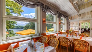 Un comedor con mesas dispuestas para cuatro, ventanas amplias con cortinas y vista a un lago.