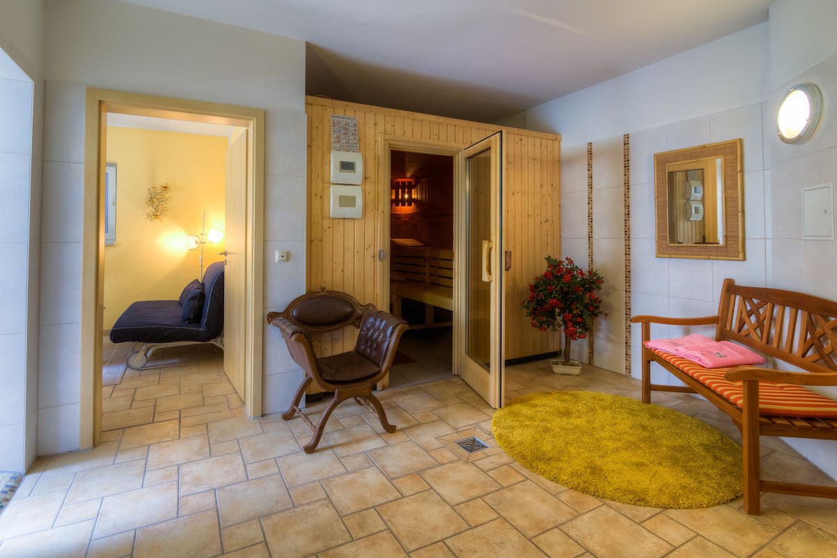 A room with a bed, a chair, a bench, and a yellow rug. There is a wooden door leading to another room.