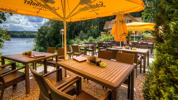 Un espacio de comedor al aire libre con mobiliario de mimbre y sombrillas amarillas, con vistas a un lago.