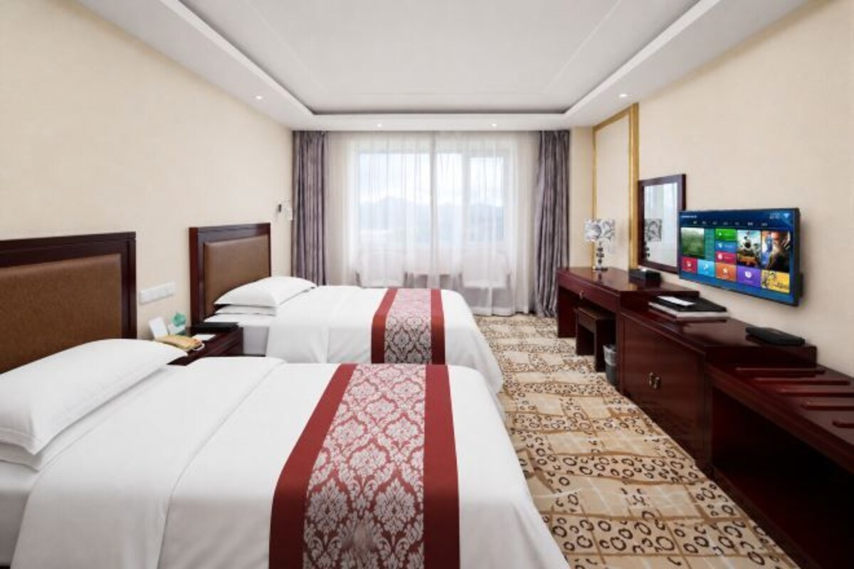 A hotel room with two beds, a television, a desk, and a large window with curtains.