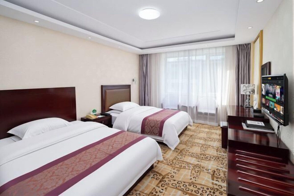 A hotel room with two beds, a television, a desk, and a large window with curtains.