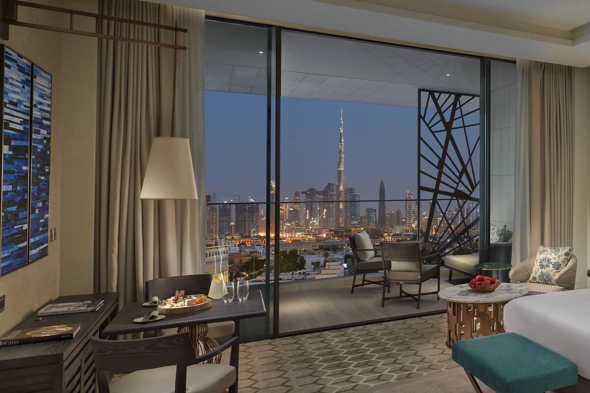 A hotel room with a large window offering a cityscape view, including a prominent tower. The room includes a bed, a desk, a chair, and a small table with a bowl of fruit.