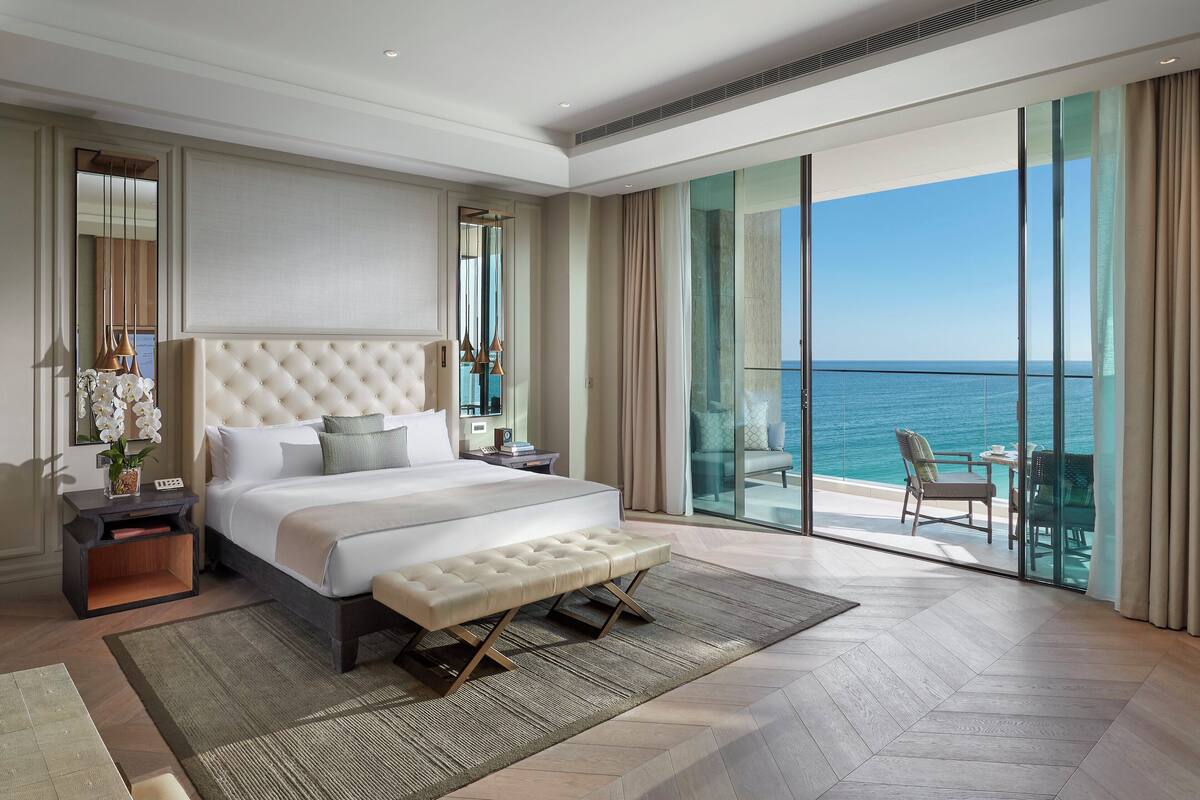 A spacious bedroom with a large bed, a bench, a nightstand, and a view of the ocean through floor-to-ceiling windows.