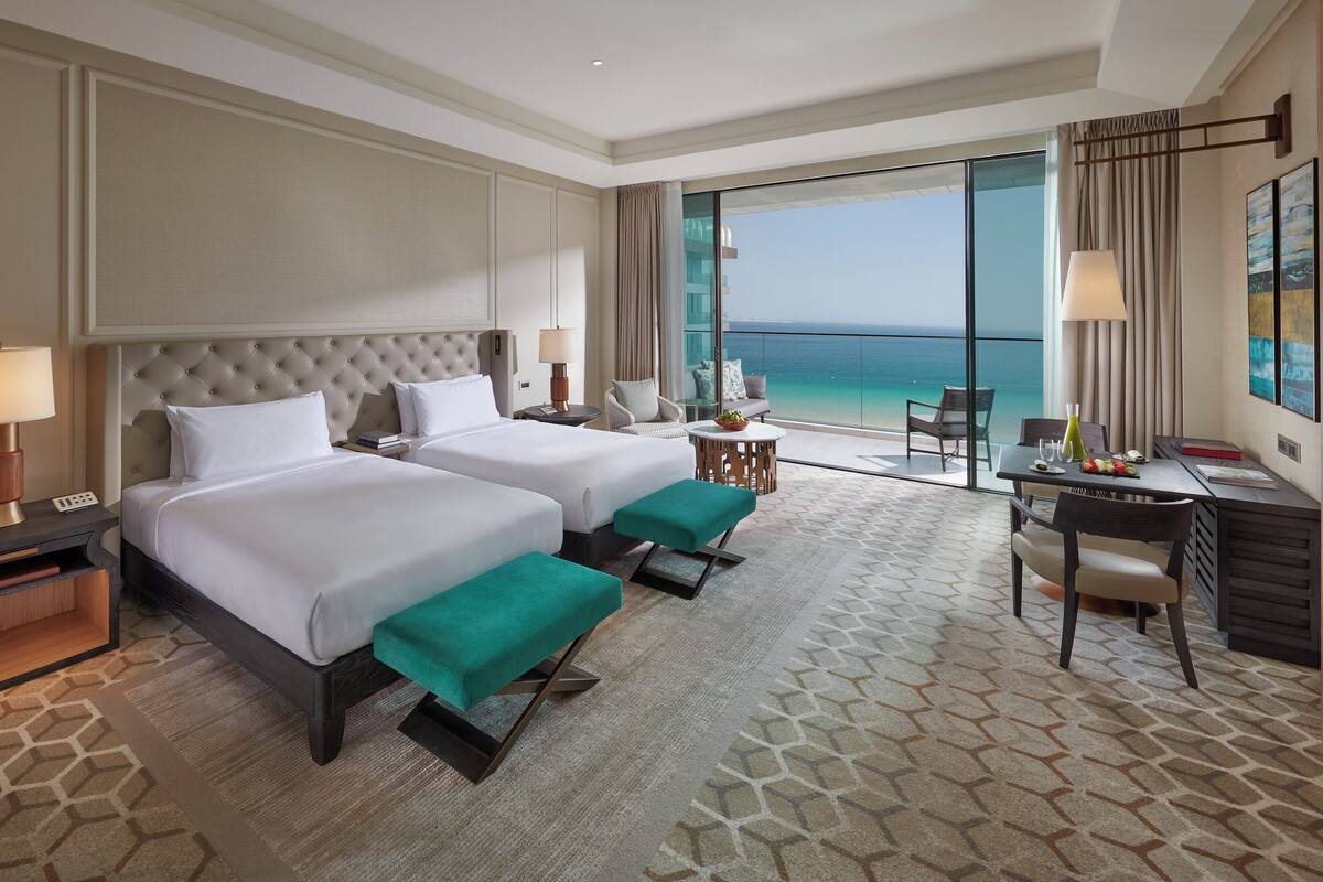 A hotel room with two beds, a desk, a chair, a small table with a plant, and a view of the ocean.
