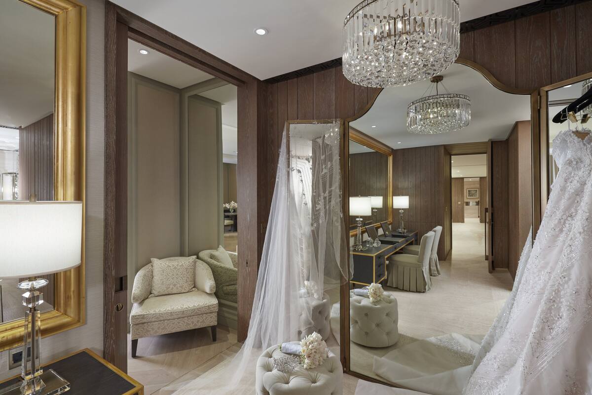 A room with a large mirror, a chandelier, a vanity with a lamp, a sofa, and a wedding dress.