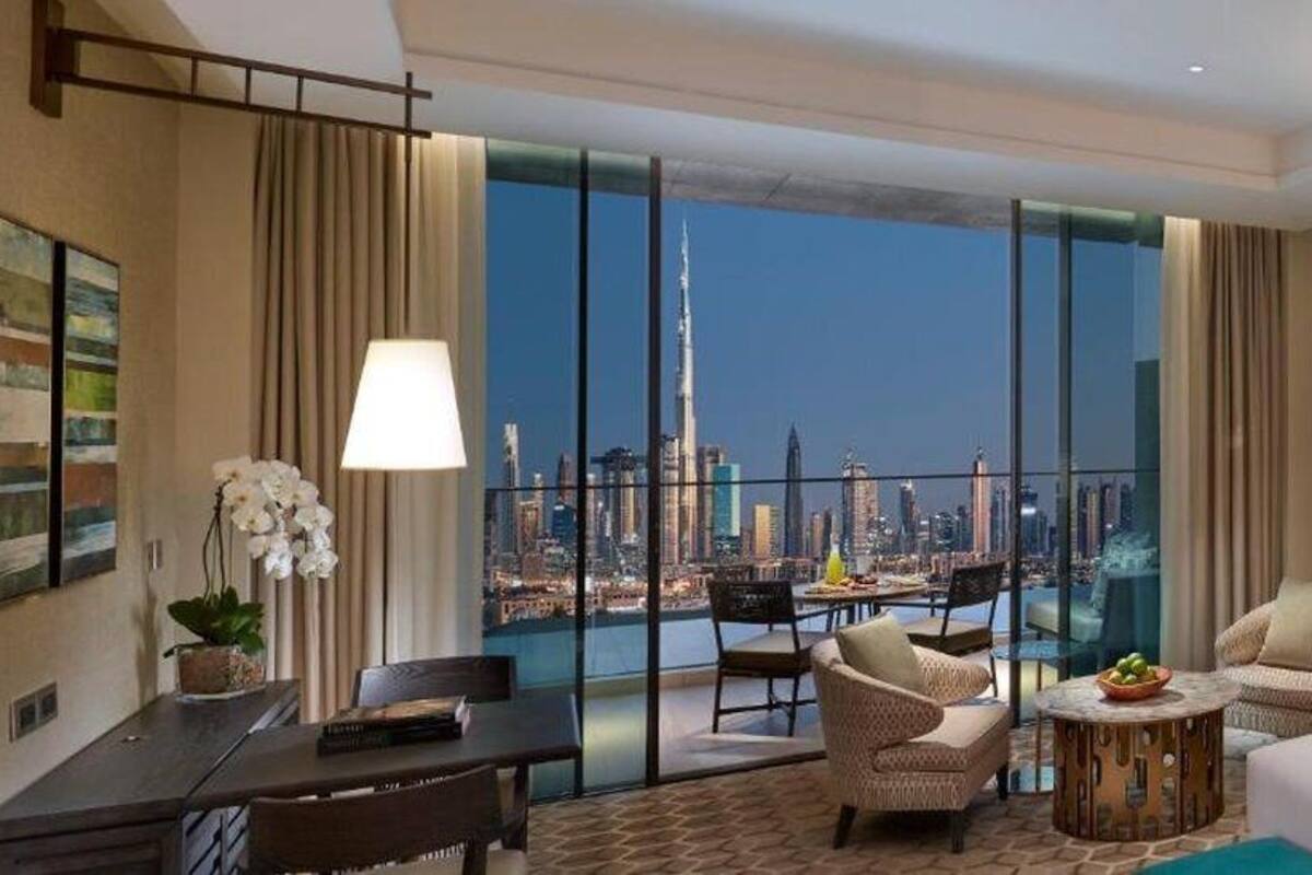 A hotel room with a large window offering a city view, a bed, two chairs, a small table, and a desk with a lamp.
