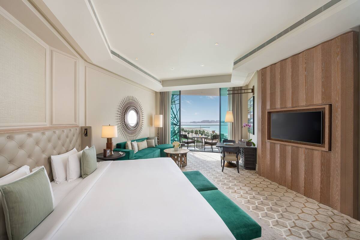 A hotel room with a large bed, a desk, and a TV. There is a view of the ocean through large windows.