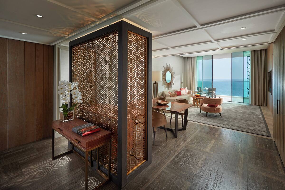 A modern hotel room with a wooden floor, a desk with a vase of flowers, a sofa, and a view of the sea through large windows.