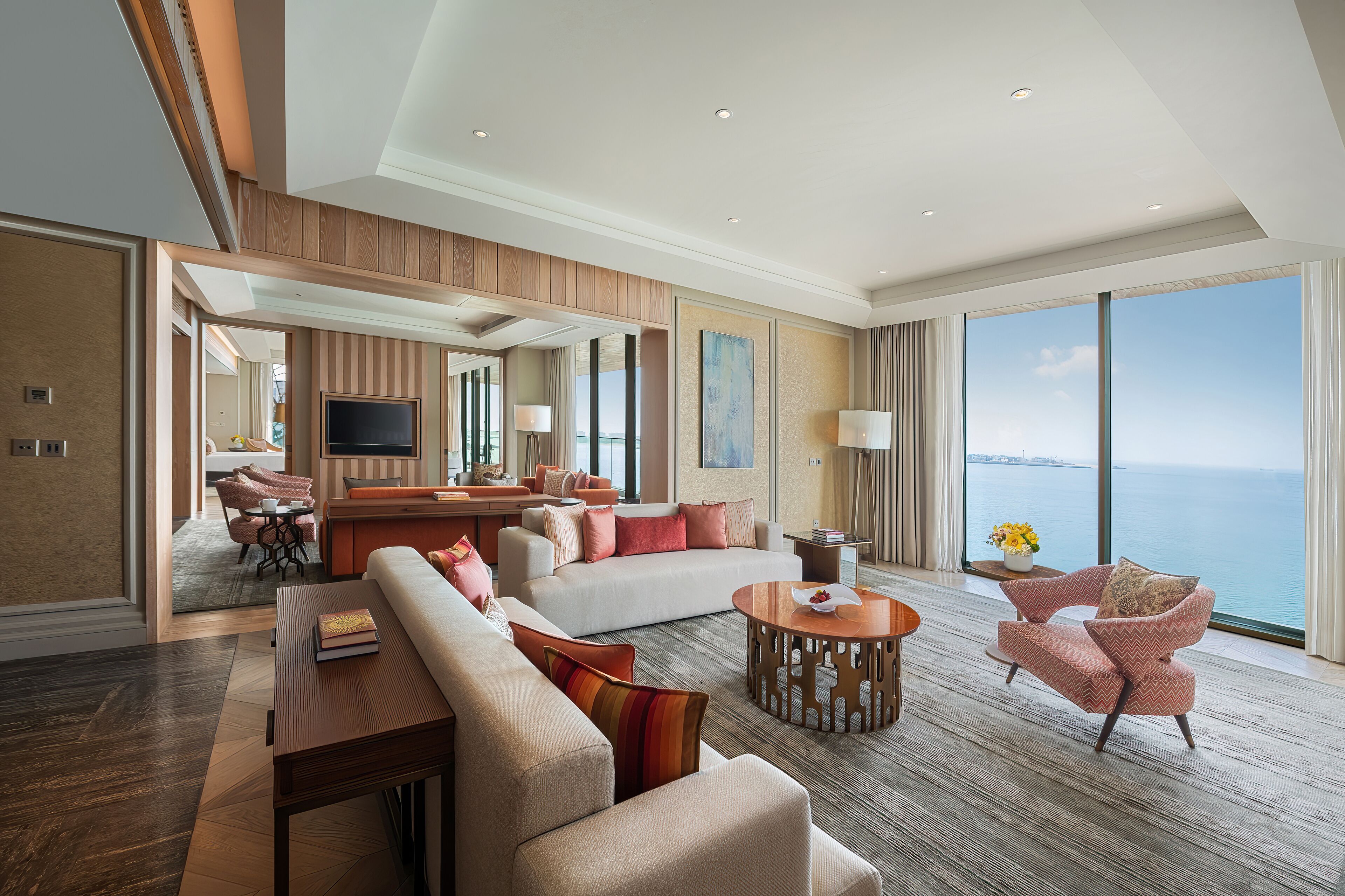 A spacious living room with a large sofa, armchairs, and a coffee table. There is a television and a dining area visible in the background.
