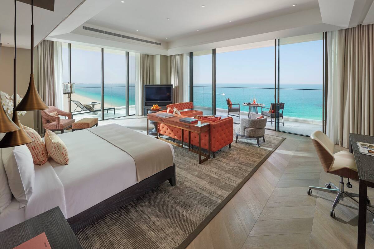 A spacious hotel room with a large bed, a seating area with a TV, and a balcony with a view of the sea.