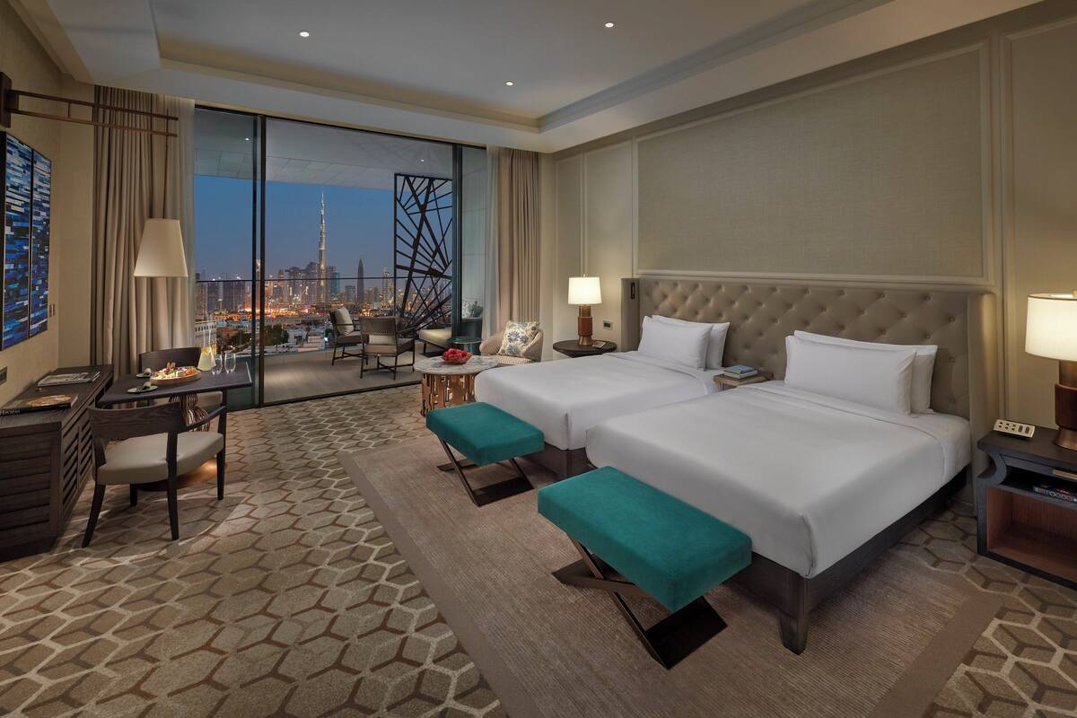 A hotel room with two beds, a desk, a chair, a table with a lamp, and a view of a cityscape.