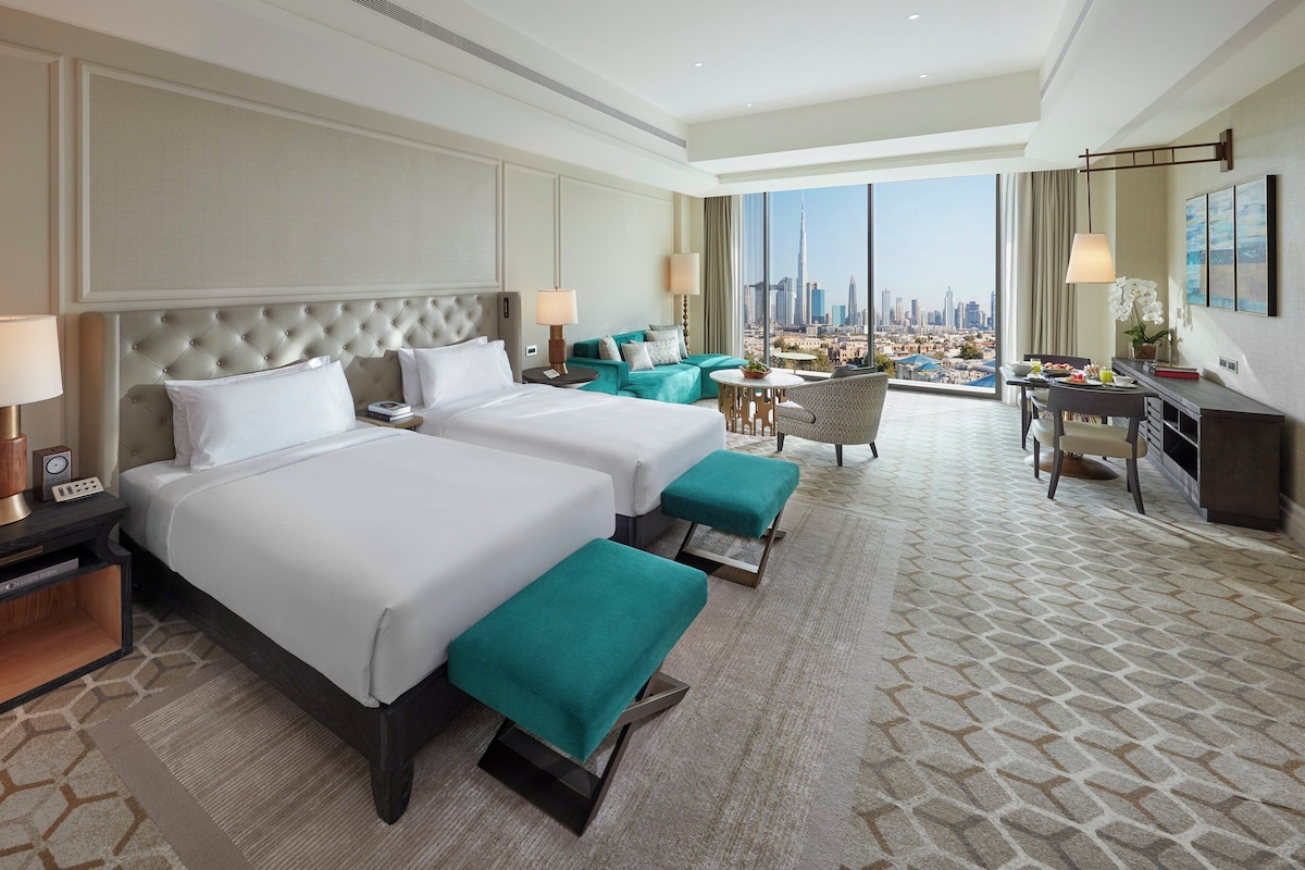 A hotel room with two beds, a desk, a chair, and a view of a city skyline.