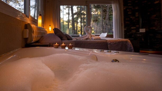 A room with a large bathtub filled with bubbles, a sofa with a blanket, and a window with a view of trees. - Mizu Hotel Boutique (Mar de Las Pampas)