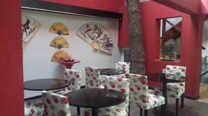 A room with a red wall, multiple round tables, and chairs with a leaf pattern. There are decorative fans on the wall and a tree trunk in the corner. - Mizu Hotel Boutique (Mar de Las Pampas)